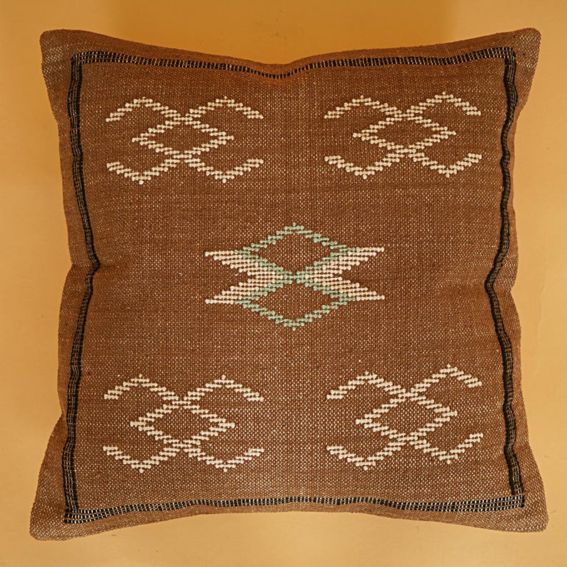 May include: Brown square pillow with a woven texture and geometric patterns. The pillow features white and teal diamond and cross-like designs. The edges are outlined with a black border, creating a decorative accent.