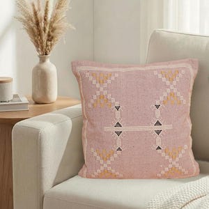 May include: A square, pink cushion with a geometric pattern in yellow, white, and black. The cushion is on a cream-coloured sofa, with a vase of dried flowers and a side table in the background.