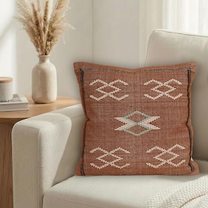 May include: A square, rust-colored decorative pillow with a geometric pattern in white and teal. The pillow is placed on a cream-colored sofa. The pillow has a black border and is part of a home decor collection.