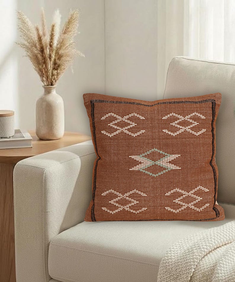 May include: A square, rust-colored decorative pillow with a geometric pattern in white and teal. The pillow is placed on a cream-colored sofa. The pillow has a black border and is part of a home decor collection.