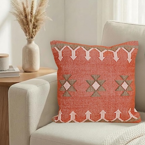 May include: A square, burnt orange decorative pillow with a geometric pattern in white and brown. The pillow is placed on a light-colored sofa, with a vase of dried flowers visible in the background. The pillow measures approximately 18 inches square.