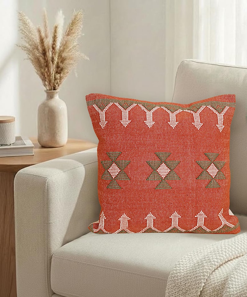 May include: A square, burnt orange decorative pillow with a geometric pattern in white and brown. The pillow is placed on a light-colored sofa, with a vase of dried flowers visible in the background. The pillow measures approximately 18 inches square.