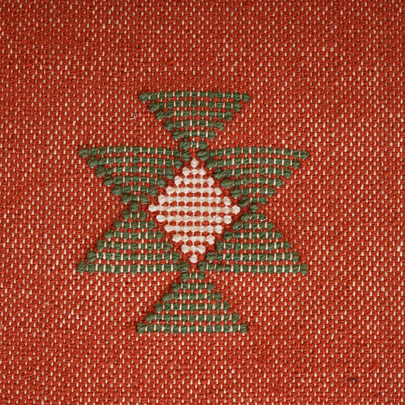 May include: Close-up of a woven textile with a repeating geometric pattern. The background is a reddish-orange color, and the pattern is composed of green and white diamond shapes.