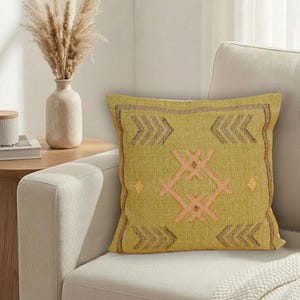 May include: A square, olive-green decorative pillow with a geometric design in peach and brown. The pillow is on a cream-colored sofa, with a vase of dried flowers and a side table in the background. The pillow has a textured appearance.