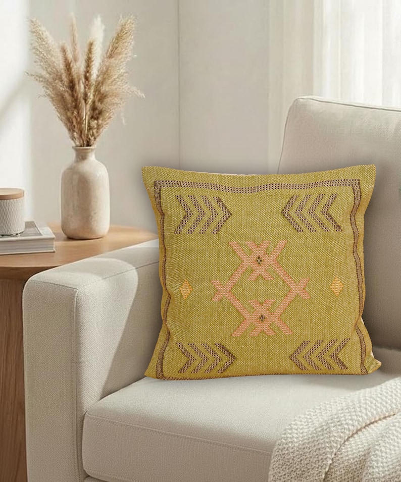May include: A square, olive-green decorative pillow with a geometric design in peach and brown. The pillow is on a cream-colored sofa, with a vase of dried flowers and a side table in the background. The pillow has a textured appearance.