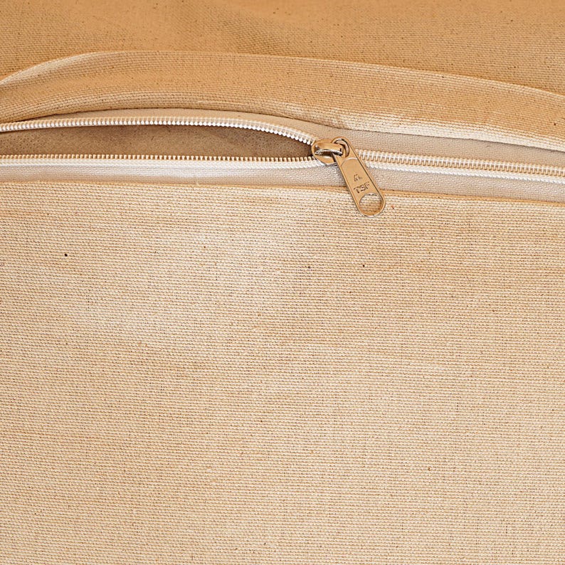May include: Close-up of a beige canvas fabric with a white zipper. The zipper is open, revealing the interior of the fabric. The zipper pull is gold-colored.