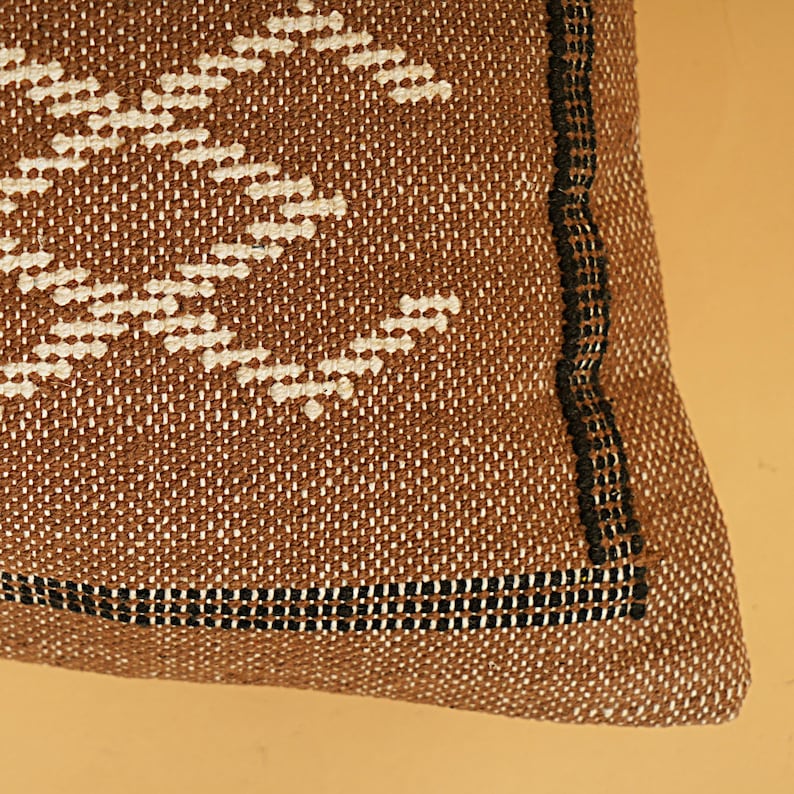 May include: Close-up of a brown woven pillow with a geometric white pattern. The pillow has a black border and a textured appearance. The design is a repeating pattern of white shapes on a brown background.