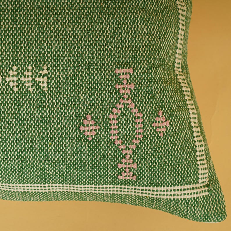 May include: Close-up of a green woven pillow with white and pink embroidered details. The pillow features a geometric pattern in pink and white stitching, with a white border. The texture of the fabric is visible.