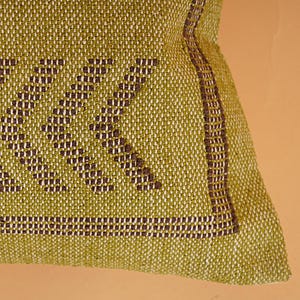 May include: Close-up of a woven pillow with a geometric pattern. The pillow features a light green background with a dark brown geometric design. A dark brown border frames the edge of the pillow.
