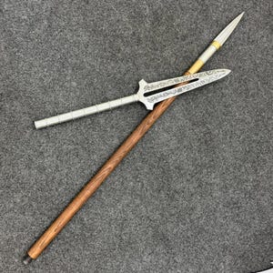 Peut inclure: Un long manche en bois avec une finition grainée brune et une extrémité métallique filetée. Une pointe de lance argentée avec un motif gravé complexe et une découpe centrale est fixée au manche, dotée d'une section de poignée nervurée.