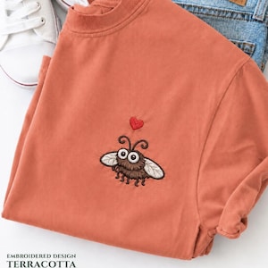 Könnte beinhalten: Terracottafarbenes T-Shirt mit einer gestickten Cartoon-Fliege und einem roten Herz. Das Shirt wird mit blauen Jeans und weißen Turnschuhen kombiniert. Der Text "EMBROIDERED DESIGN TERRACOTTA" befindet sich unten.