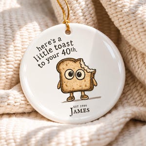 May include: A round white ceramic ornament with a gold hanging string. The design features a cartoon piece of toast with a bite taken out of it, the text "here's a little toast to your 40th.", and the text "EST 1986 JAMES" printed in black.