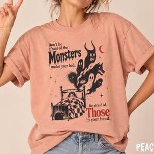 May include: A peach-colored t-shirt featuring a black and red graphic print of a bed with a person under a checkered blanket, surrounded by various monster illustrations and the text: Don't be afraid of the MONSTERS under your bed. Be afraid of Those in your head.