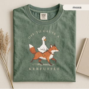 Könnte beinhalten: Moosgrünes T-Shirt mit einer Grafik einer weißen Gans, die auf einem Fuchs reitet. Der Text "OFF TO CAUSE A KERFUFFLE" ist oberhalb und unterhalb der Illustration aufgedruckt. Das Shirt hat einen Rundhalsausschnitt und kurze Ärmel.