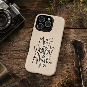 May include: A cream-coloured smartphone case featuring the black text "Me? Weird? Always." written in a stylised, swirling font. Below the text is a small, simple line drawing of a house. The case has a black camera cutout for three lenses.