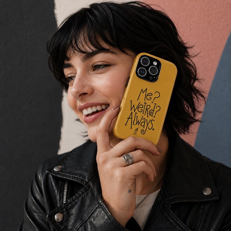 May include: A mustard yellow smartphone case featuring the black printed text Me? Weird? Always. with a small graphic of a house underneath. The case has a cutout for a multi-lens camera system and covers the back and sides of the mobile device.