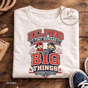 Könnte beinhalten: Elfenbeinfarbenes T-Shirt mit einem rot-weiß-blauen Grafikdesign. Das Design zeigt den Text "HELPING TINY HUMANS DO BIG THINGS SHAPING FUTURES" mit Illustrationen von zwei Kindern. Blaue Leinenschuhe sind sichtbar.
