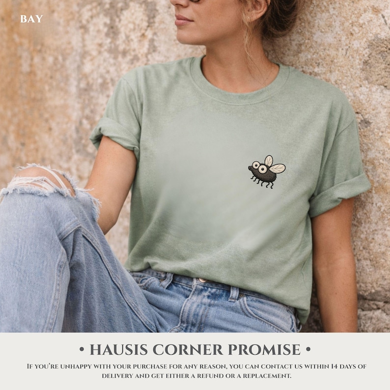 May include: Sage green t-shirt featuring a cartoon fly graphic. The shirt has rolled-up sleeves and is paired with blue jeans. The word "BAY" is visible in the top left corner.