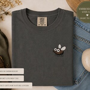 May include: A dark gray Comfort Colors tee shirt with a small embroidered fly design. The fly is brown with white wings and large eyes. The shirt has a crew neck and a tag that reads "Comfort Colors".