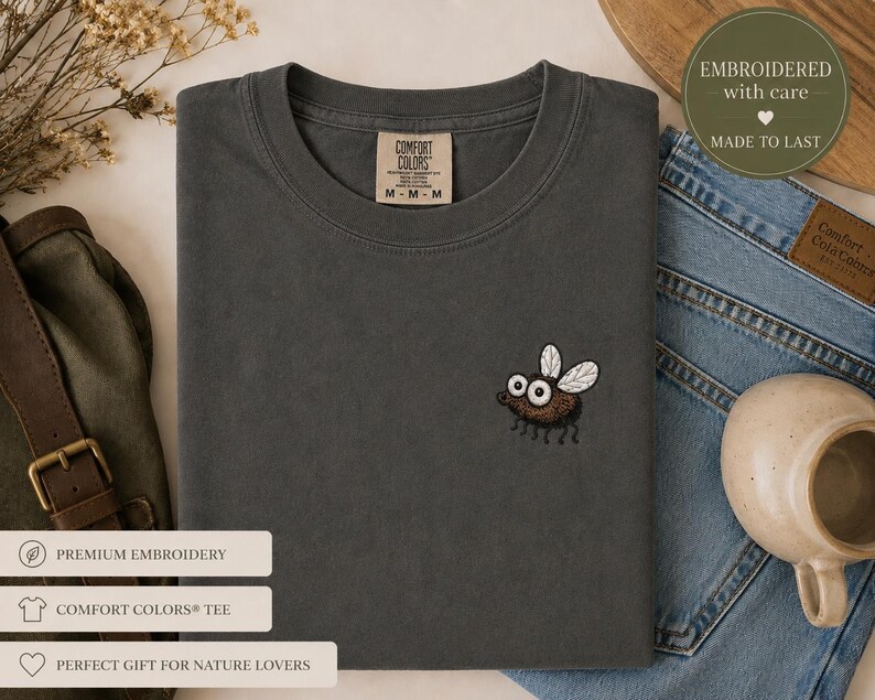 May include: A dark gray Comfort Colors tee shirt with a small embroidered fly design. The fly is brown with white wings and large eyes. The shirt has a crew neck and a tag that reads "Comfort Colors".