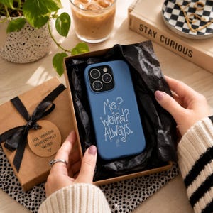 May include: A blue smartphone case featuring the white text "Me? WEIRD? Always." and a small house icon, nestled inside a gift box lined with black tissue paper, presented as a gift with a tag reading "FOR SOMEONE WONDERFULLY WEIRD."