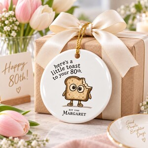 May include: A round white ceramic ornament featuring a cartoon slice of toast with large eyes and brown shoes. The text reads: here's a little toast to your 80th. EST 1946 MARGARET. The ornament hangs from a gold cord tied with a cream satin ribbon.