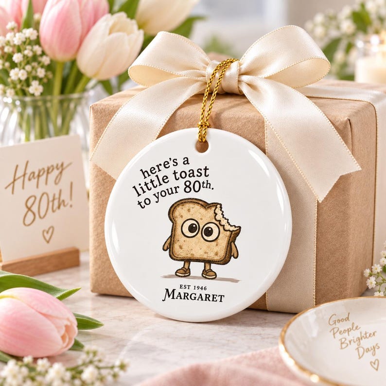 May include: A round white ceramic ornament featuring a cartoon slice of toast with large eyes and brown shoes. The text reads: here's a little toast to your 80th. EST 1946 MARGARET. The ornament hangs from a gold cord tied with a cream satin ribbon.