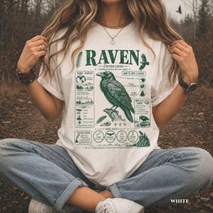 May include: White t-shirt with a detailed green graphic of a raven, scientific information, and the word "RAVEN". The design includes illustrations of the bird, its habitat, and facts. The shirt is worn by a person.