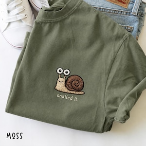 May include: An olive green t-shirt featuring an embroidered graphic of a light brown snail with large eyes and a dark brown shell. Below the snail, the text "snailed it." is embroidered in white. The shirt is paired with blue denim jeans.