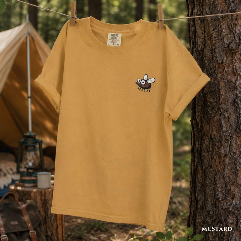 May include: Mustard-colored t-shirt with a cartoon fly graphic on the front. The short-sleeved shirt is hanging on a clothesline. The word "MUSTARD" is printed in white on the bottom right.