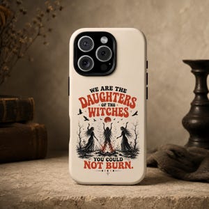 May include: A cream-colored phone case featuring a graphic illustration of three figures standing around a fire. The text reads, "WE ARE THE DAUGHTERS OF THE WITCHES YOU COULD NOT BURN." The design includes silhouettes of trees, birds, and a red moon.