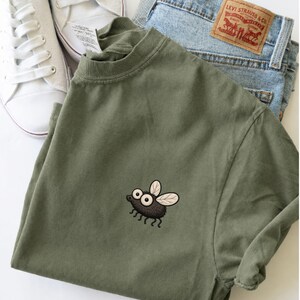 May include: An olive green t-shirt with an embroidered cartoon fly design. The shirt is paired with light blue denim jeans and white sneakers. The text "EMBROIDERED DESIGN MOSS" is at the bottom of the image.