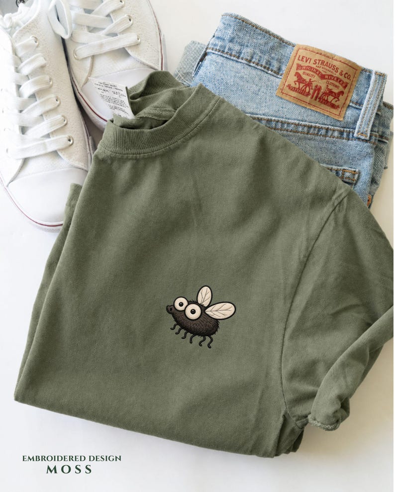 May include: An olive green t-shirt with an embroidered cartoon fly design. The shirt is paired with light blue denim jeans and white sneakers. The text "EMBROIDERED DESIGN MOSS" is at the bottom of the image.
