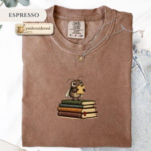 May include: Espresso-colored t-shirt with an embroidered design of a cartoon insect holding a paper, standing on a stack of books. The shirt has a Comfort Colors label and a silver necklace. The word "embroidered" is on a label.