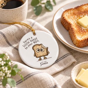 May include: A round white ceramic ornament with a gold hanging string. The design features a cartoon slice of toast with eyes and limbs, the text "here's a little toast to your 40th.", and the name "James" with "EST 1986" printed below it.