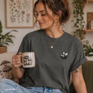 May include: Dark grey t-shirt with a small embroidered fly on the chest. The shirt is worn with blue jeans. A beige mug with a mushroom design is held in the hand. The word "PEPPER" is printed at the bottom.