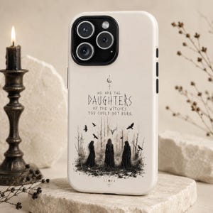 May include: A cream-colored smartphone case featuring a black graphic print of three figures in long dresses standing in a forest. The text reads, "WE ARE THE DAUGHTERS OF THE WITCHES YOU COULD NOT BURN." The design includes birds, a deer skull, and moon phases.
