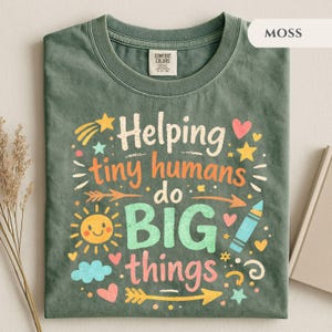 Könnte beinhalten: Moosgrünes T-Shirt mit dem farbenfrohen Schriftzug "Helping tiny humans do BIG things". Das Design umfasst eine lächelnde Sonne, Sterne, Herzen und einen Pfeil. Das Shirt ist aus weichem Material.