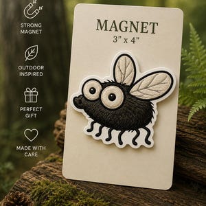 May include: A black and white fly magnet, 3" x 4", with large eyes and detailed wings, is displayed on a cream-colored card. The card has the word "MAGNET" printed at the top. The magnet is on a wooden surface.
