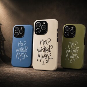 May include: Three smartphone cases in blue, cream, and olive green. Each case features a black camera module and the printed text "Me? weird? Always." in a stylised font, accompanied by a small illustration of a house at the bottom.
