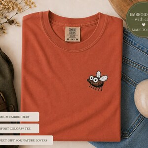 May include: Rust-colored Comfort Colors tee shirt with an embroidered cartoon fly. The shirt is folded and placed on a white surface, next to a pair of blue jeans and a ceramic mug. The shirt has the text "COMFORT COLORS" on the tag.