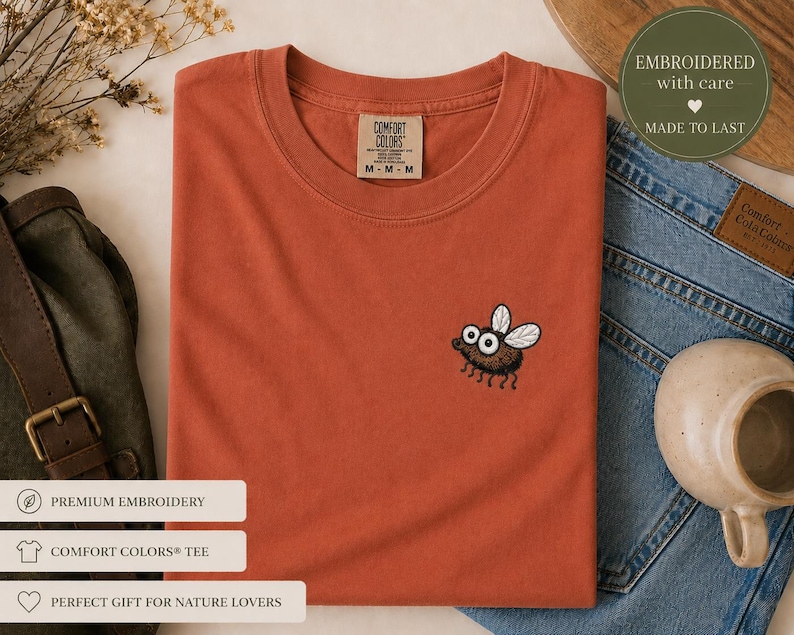 May include: Rust-colored Comfort Colors tee shirt with an embroidered cartoon fly. The shirt is folded and placed on a white surface, next to a pair of blue jeans and a ceramic mug. The shirt has the text "COMFORT COLORS" on the tag.