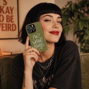 May include: An olive green smartphone case featuring a triple camera cutout and the white printed text "Me? WEIRD? Always." in a stylised, handwritten font. The case has a smooth matte finish and covers the back of the mobile device.