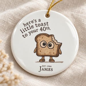 May include: A round white ceramic ornament with a gold hanging string. It features a cartoon slice of toast with large eyes and a bite mark, with the text "here's a little toast to your 40th." and "EST 1986 JAMES" printed in dark brown ink.