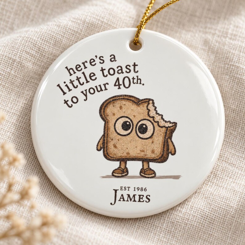 May include: A round white ceramic ornament with a gold hanging string. It features a cartoon slice of toast with large eyes and a bite mark, with the text "here's a little toast to your 40th." and "EST 1986 JAMES" printed in dark brown ink.