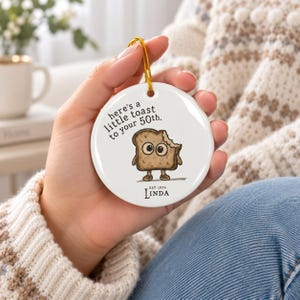 May include: A round white ceramic ornament featuring a cartoon slice of toast with a bite taken out of it. The text reads, "here's a little toast to your 50th." and "EST 1976 LINDA" at the bottom. It includes a gold hanging string at the top.