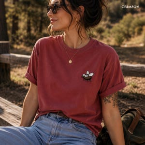 May include: Crimson-colored t-shirt with a cartoon fly graphic. The short-sleeved shirt has rolled-up sleeves and is made of a soft material. The word "CRIMSON" is printed in the upper right corner.