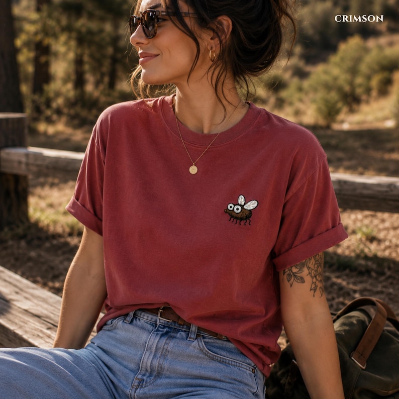 May include: Crimson-colored t-shirt with a cartoon fly graphic. The short-sleeved shirt has rolled-up sleeves and is made of a soft material. The word "CRIMSON" is printed in the upper right corner.