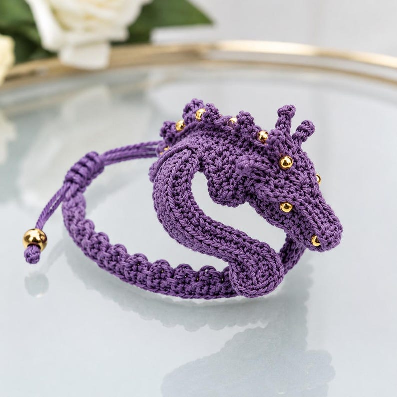 May include: A purple crocheted bracelet featuring a seahorse-shaped design. The seahorse is adorned with small, round, metallic gold beads along its back and head. The bracelet includes an adjustable cord with a gold bead at the end of the string.