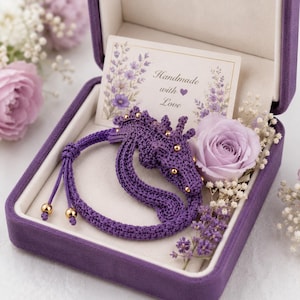 May include: A purple crocheted dragon bracelet with small gold beads, presented in a purple velvet box alongside a light purple rose, dried flowers, and a card with the text Handmade with Love, featuring a small heart icon near the text.